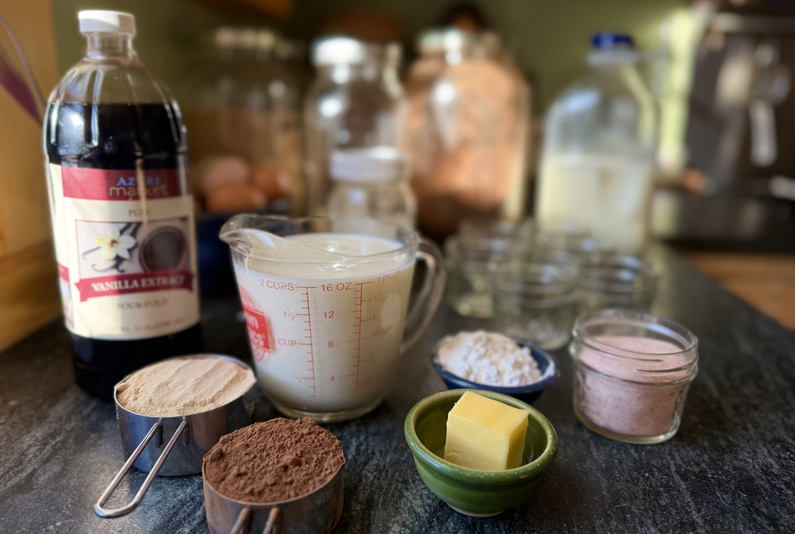 Measured ingredients for homemade chocolate pudding including milk, cocoa powder, maple sugar, cornstarch, vanilla extract, butter, and salt.