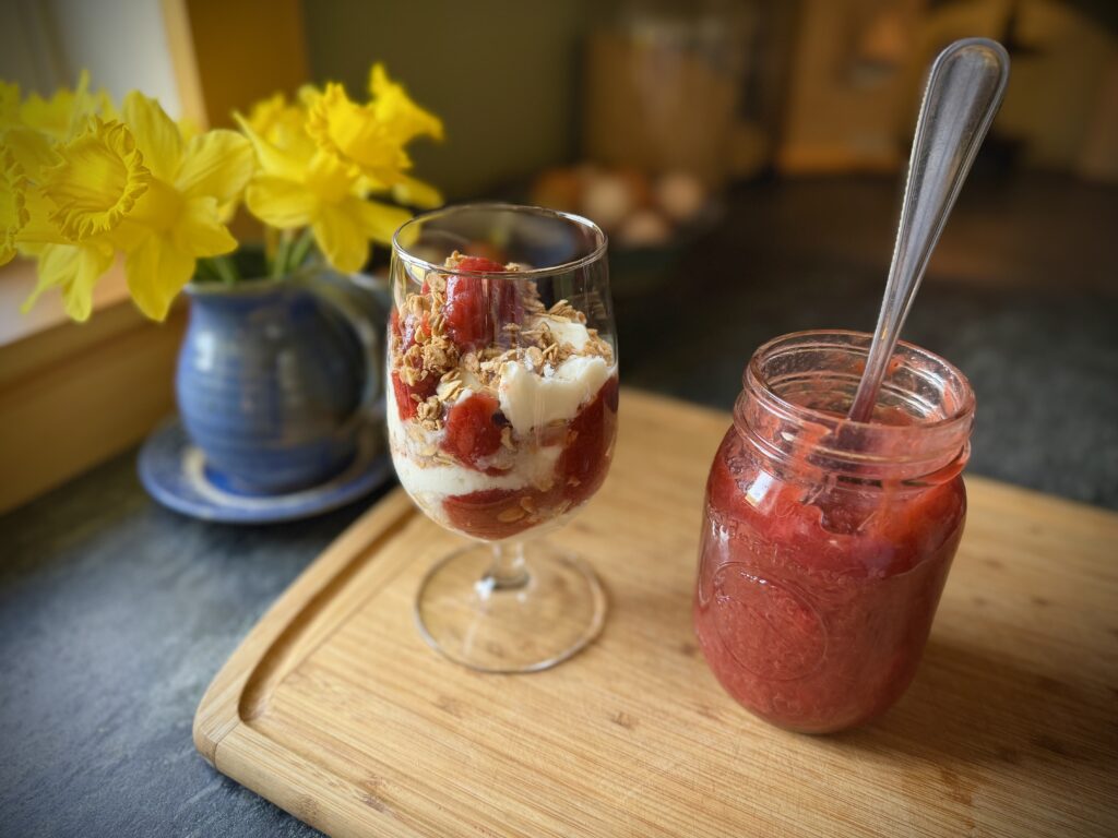 Strawberry rhubarb topping layered in a yogurt parfait with granola in a glass, served with a jar of homemade topping