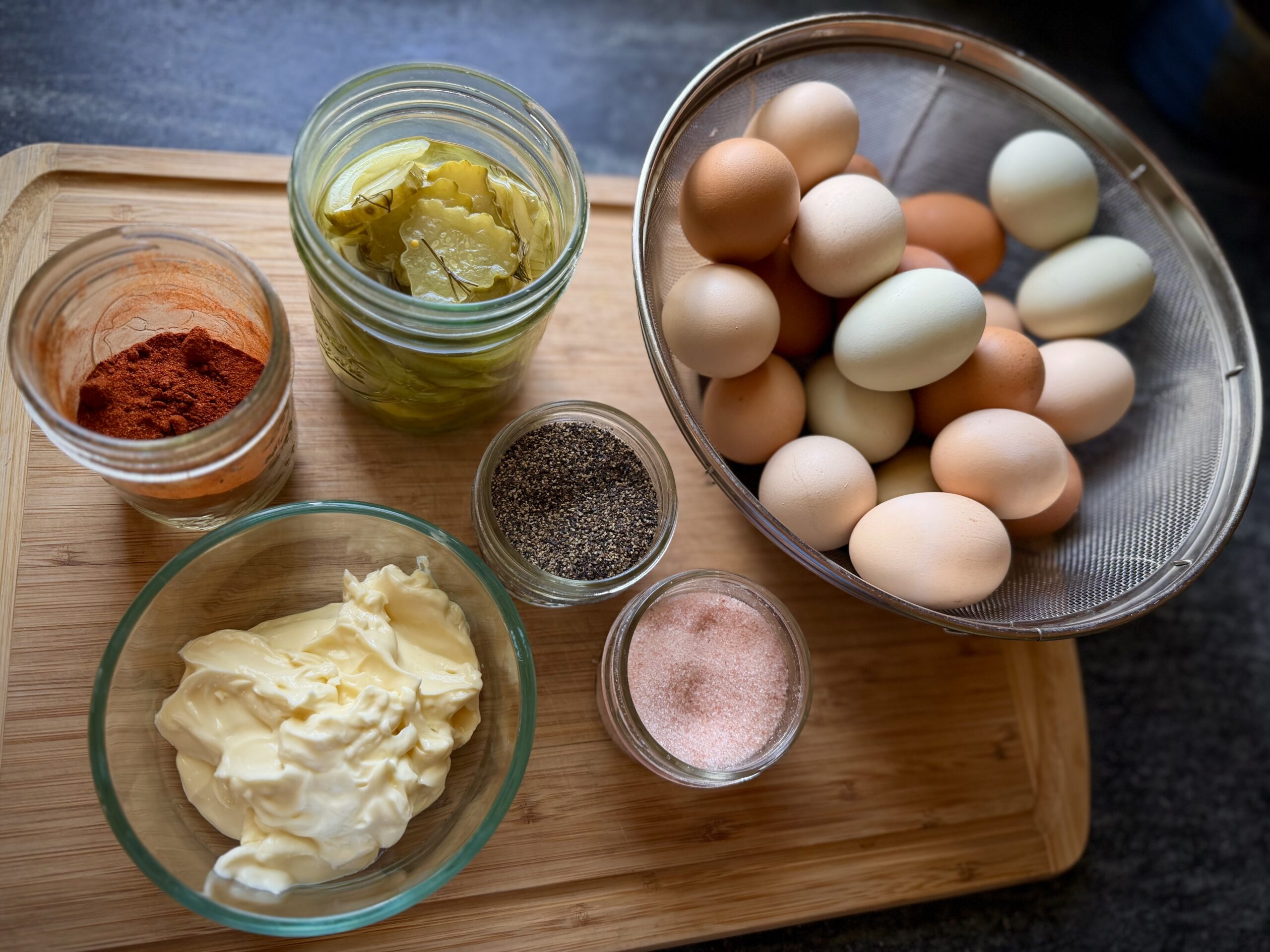 Farm fresh eggs, homemade mayonnaise, paprika, salt, pepper, and sliced pickles arranged on a wooden board for making simple homemade deviled eggs.
