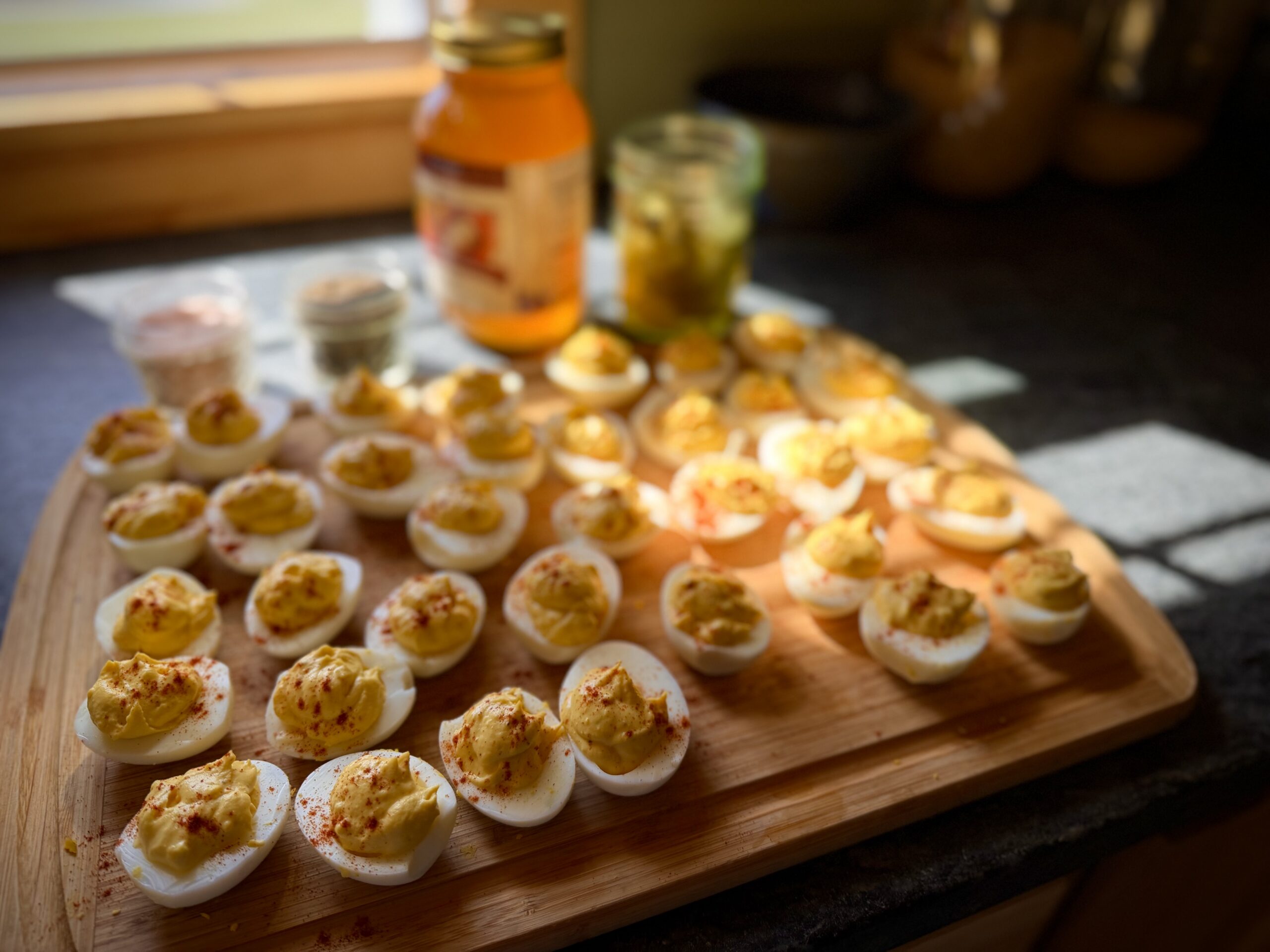 Simple homemade deviled eggs sprinkled with paprika and arranged on a wooden board in warm afternoon light beside jars of pickles and seasonings.