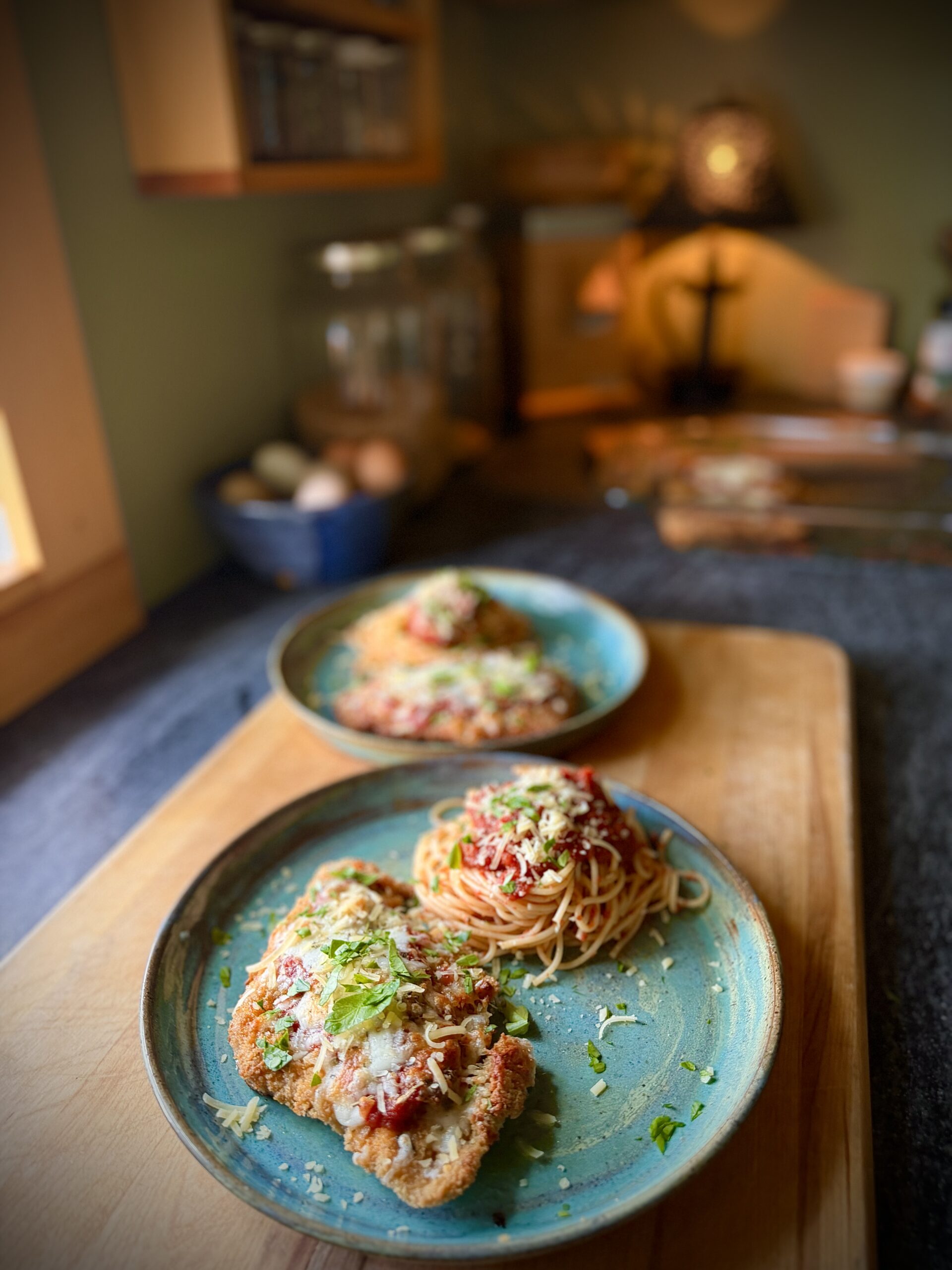 Homemade chicken parmesan served with spaghetti, marinara sauce, parmesan cheese, and fresh basil on blue dinner plates.