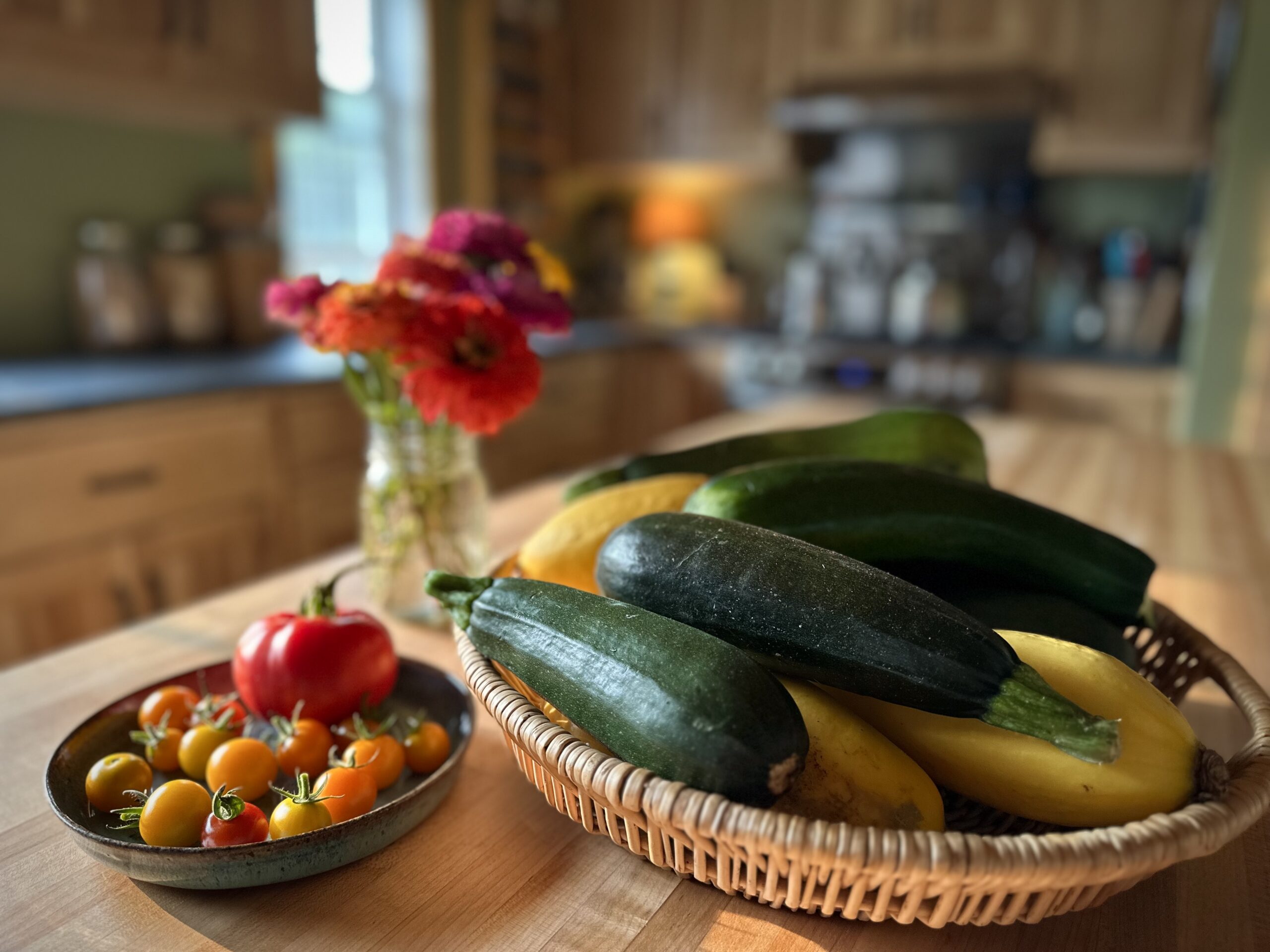 Basket of homegrown zucchini and summer squash on a farmhouse table with fresh tomatoes and garden flowers in the background.