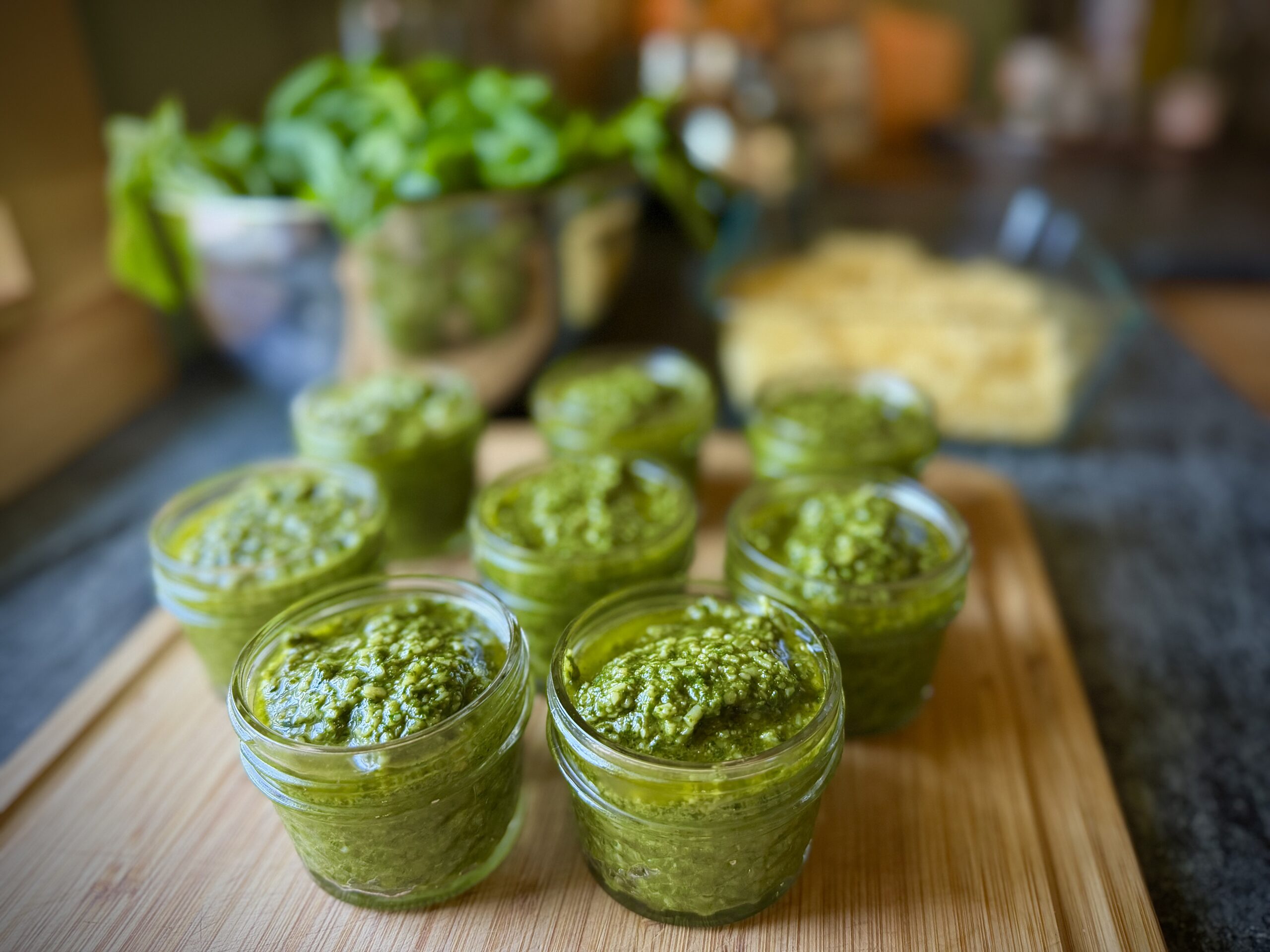 Small glass jars filled with homemade basil pesto on a wooden cutting board, with fresh basil and grated Parmesan in the background.