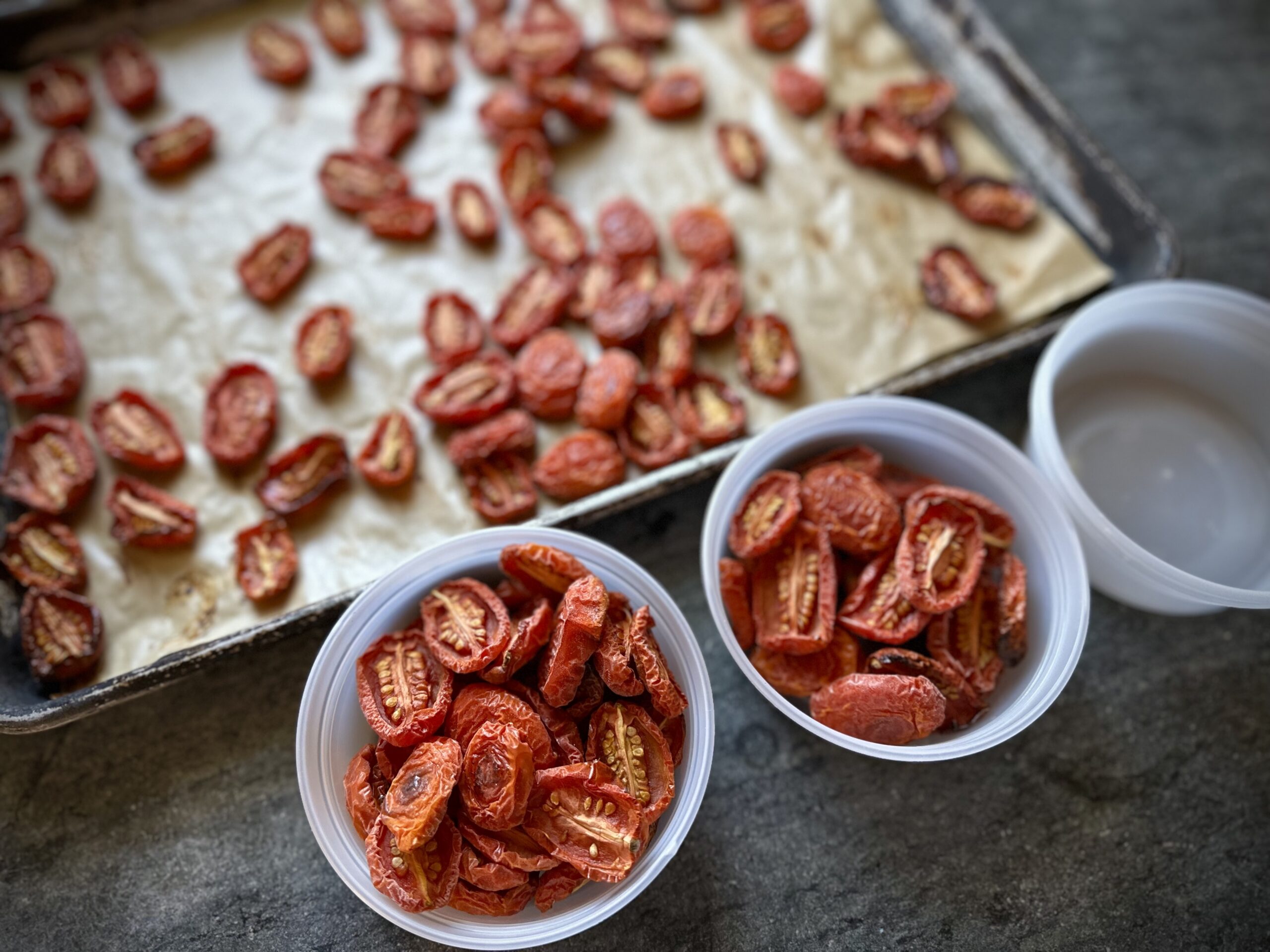 Oven-roasted Juliet tomatoes on a parchment-lined baking sheet, with two small containers filled for freezer storage.