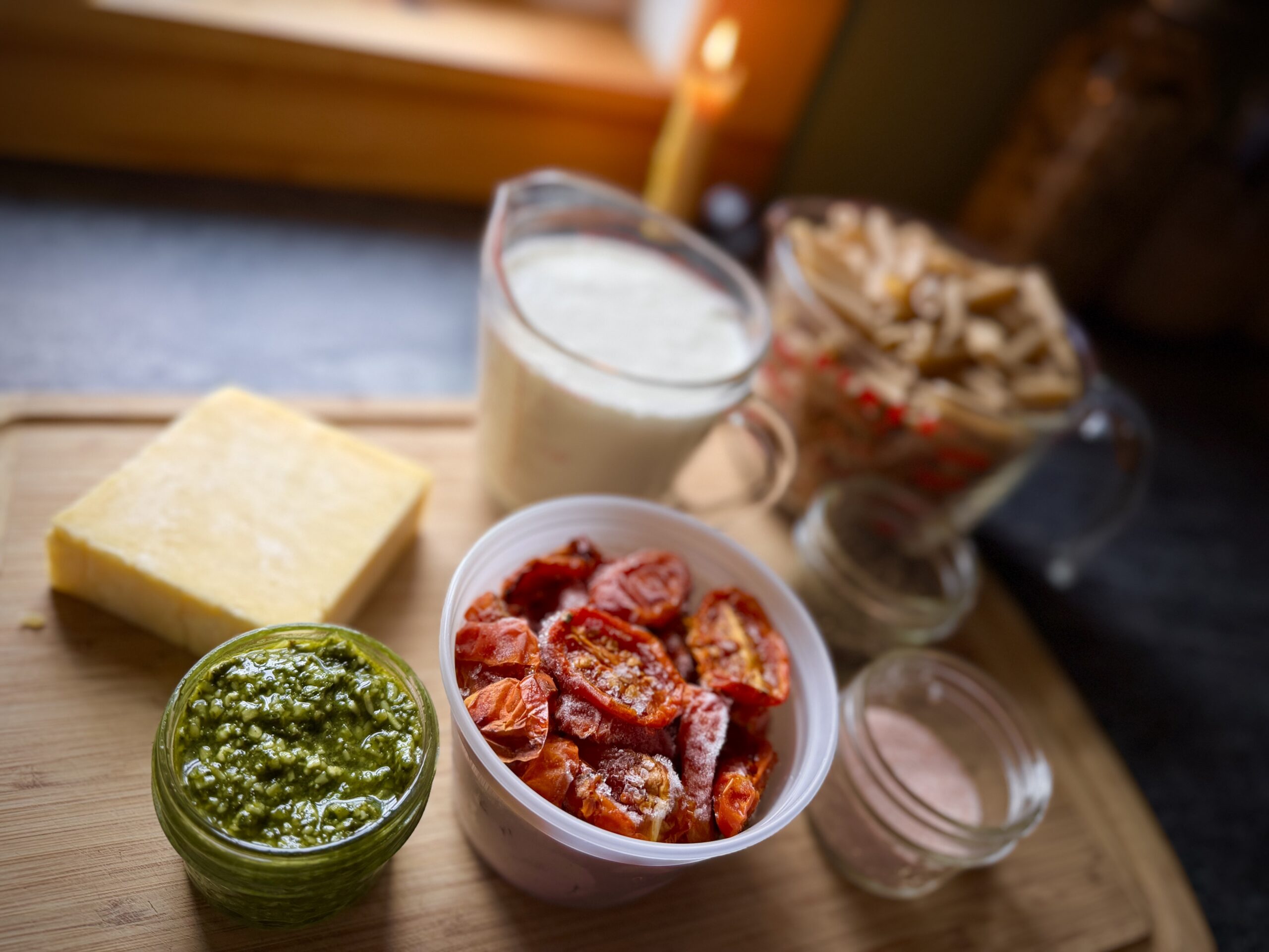 Ingredients for homemade pesto pasta arranged on a wooden cutting board, including basil pesto, oven-roasted tomatoes, Parmesan cheese, heavy cream, and pasta.