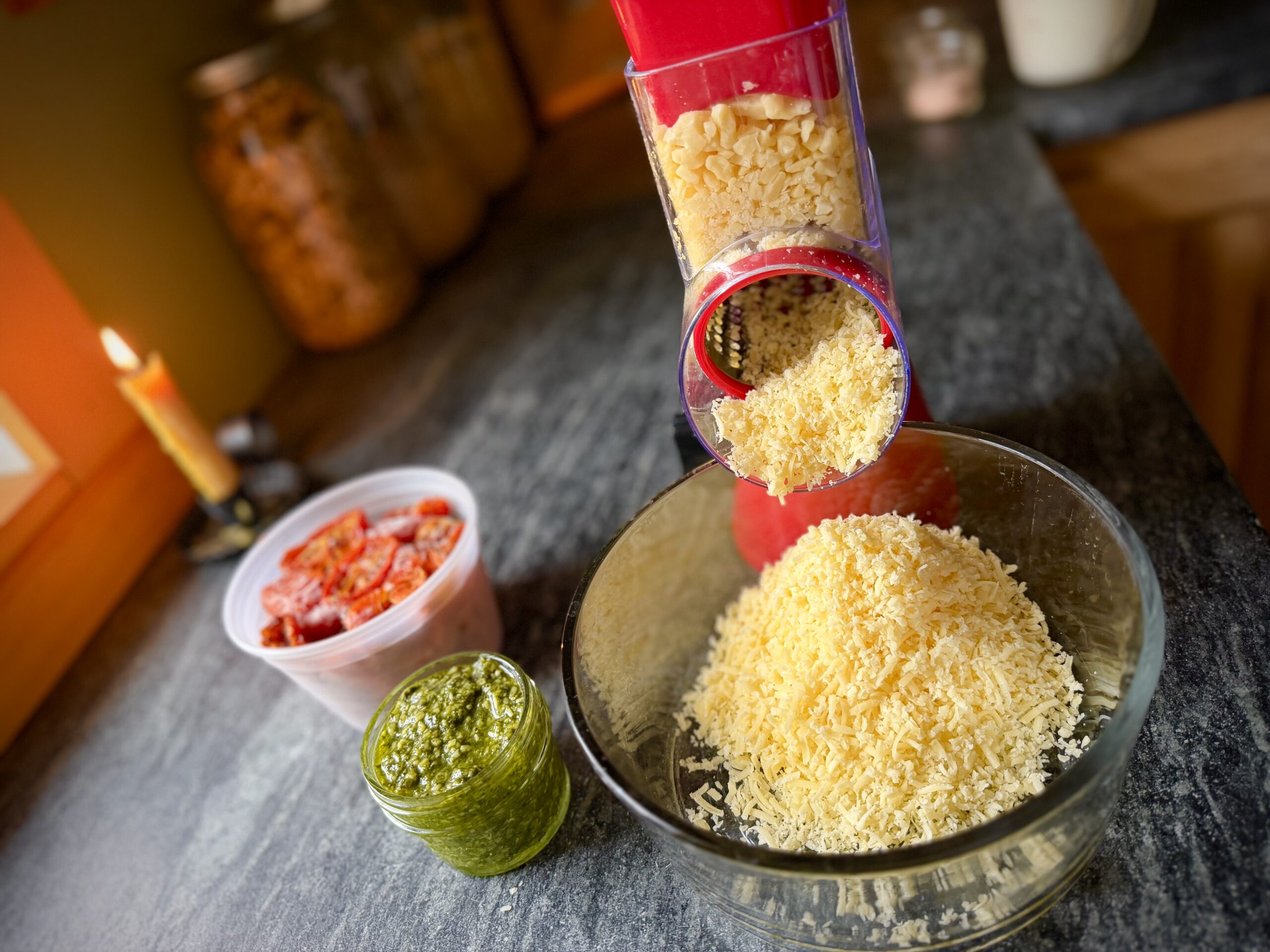 Freshly grated Parmesan cheese falling from a rotary grater into a glass bowl, with homemade basil pesto and oven-roasted tomatoes alongside.