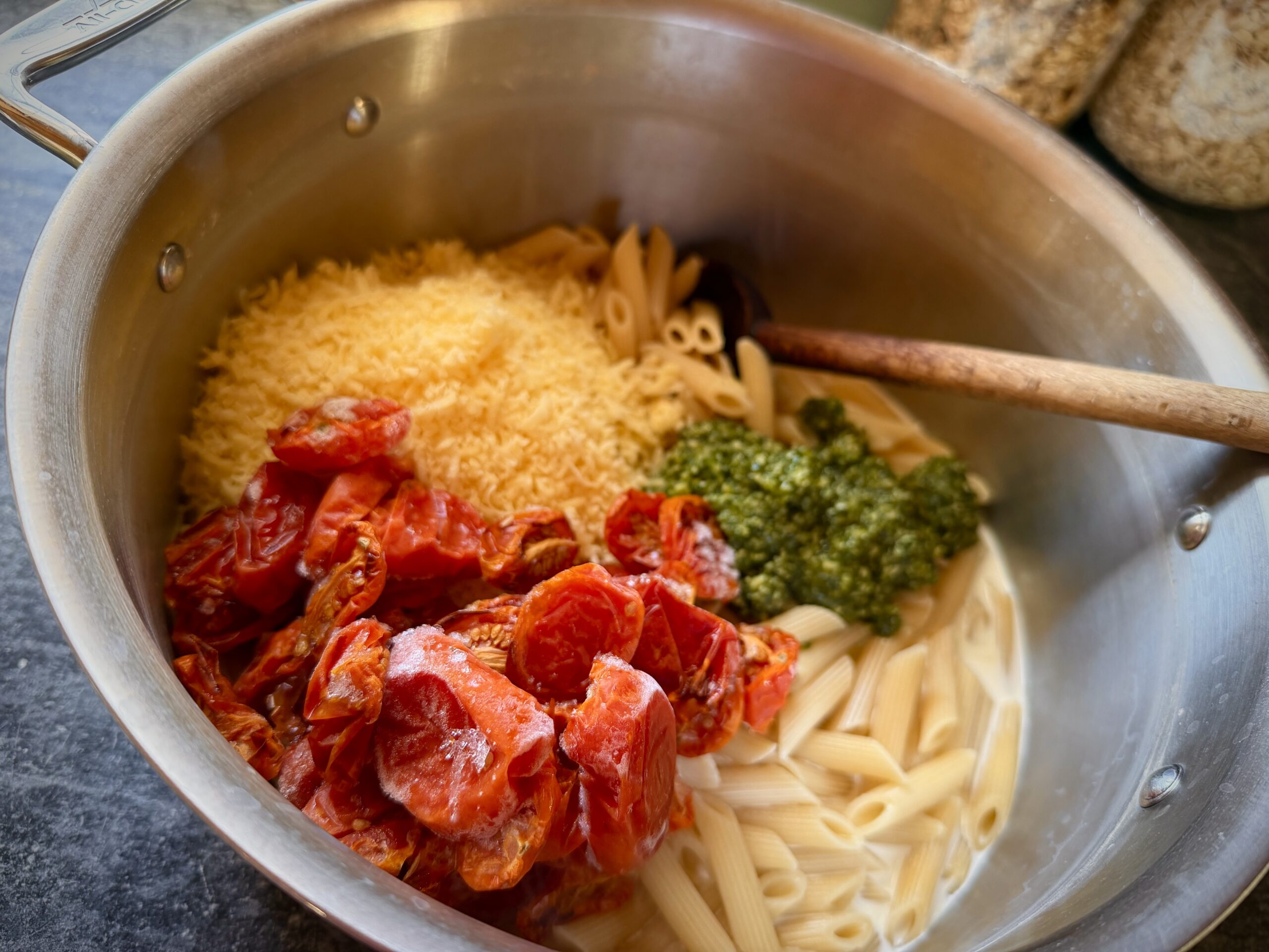 Cooked penne pasta with homemade basil pesto, oven-roasted tomatoes, freshly grated Parmesan, and cream in a stainless steel pot, ready to be stirred together.