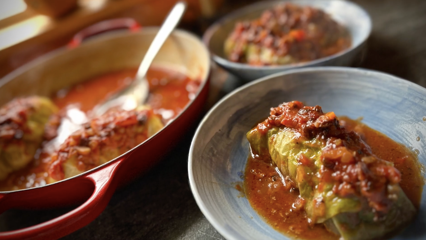Homemade stuffed cabbage roll topped with rich tomato sauce in a blue bowl, with more cabbage rolls in a red Dutch oven.