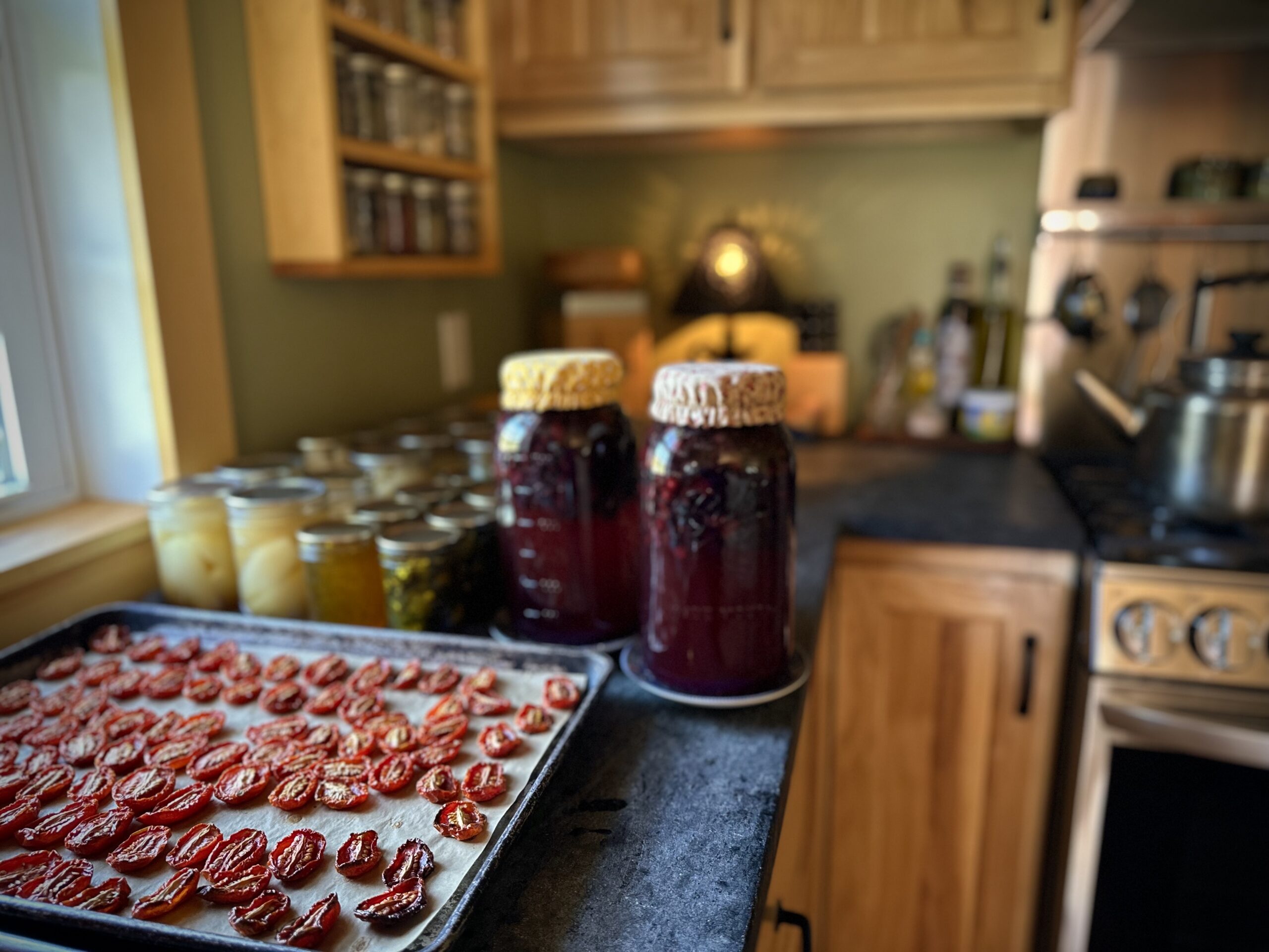 Oven-roasted Juliet tomato halves on a parchment-lined baking sheet in a farmhouse kitchen, surrounded by jars of preserved garden produce.